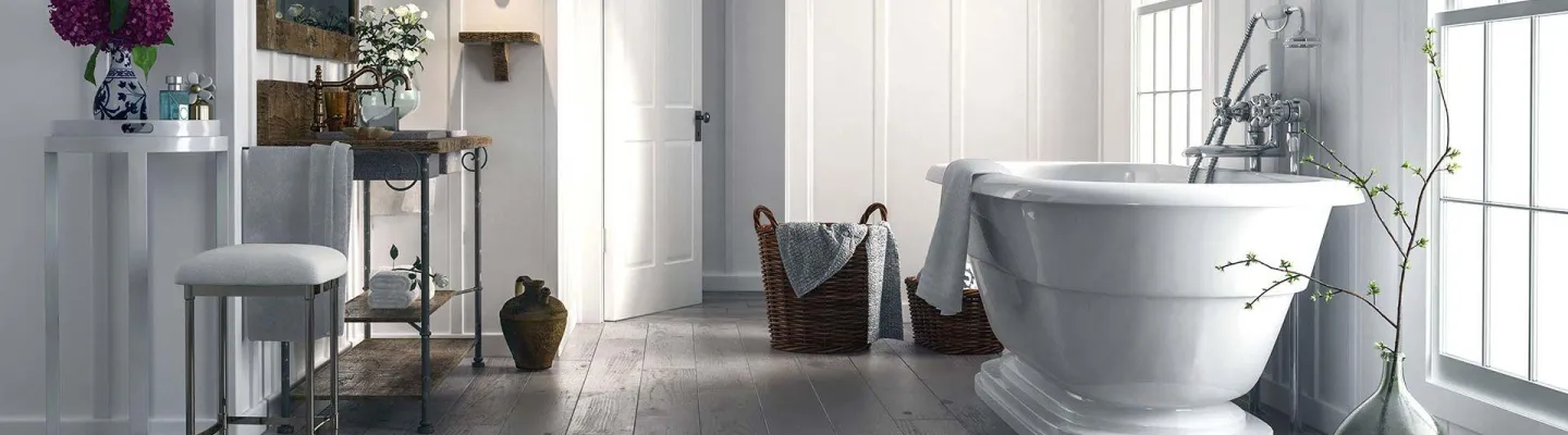 A luxurious bathroom interior featuring a freestanding white bathtub, grey wooden flooring, and bright natural light from large windows. A vanity with purple flowers and decorative items sits on the left, with wicker baskets and potted plants completing the spa-like aesthetic.