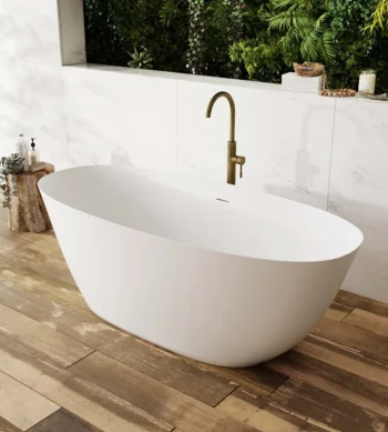 White freestanding bathtub on wooden floor with brass faucet, marble wall tiles and lush greenery visible in the background.