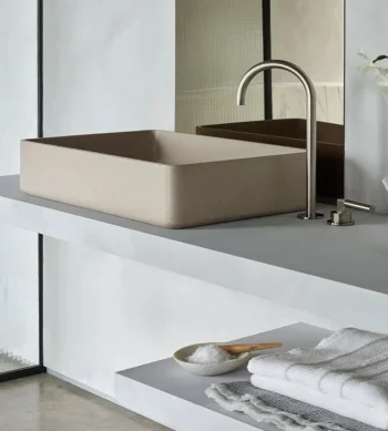 Beige rectangular vessel sink on a grey countertop with a curved chrome faucet; mirrors behind and a shelf below with white towels and a small dish.