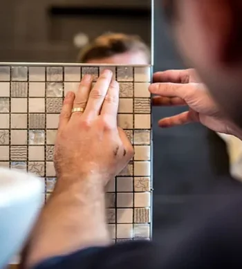 Close-up of hands placing small textured square tiles on a grid panel, with a ring on one finger and another person guiding the board.