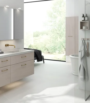 Bright modern bathroom with light-wood vanity, brass tap, large window view of greenery, wall cabinets, glass shower and towel rack.