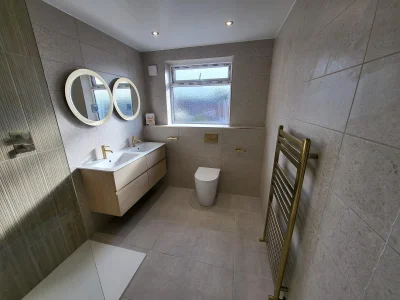 Modern grey-tiled bathroom with a wall-hung light-wood double vanity, two round backlit mirrors, brass taps, frosted window, toilet and a gold towel radiator.